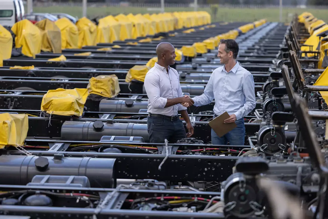 Two men shake hands amid rows of business equipment, one holding paperwork, yellow tarps visible in background