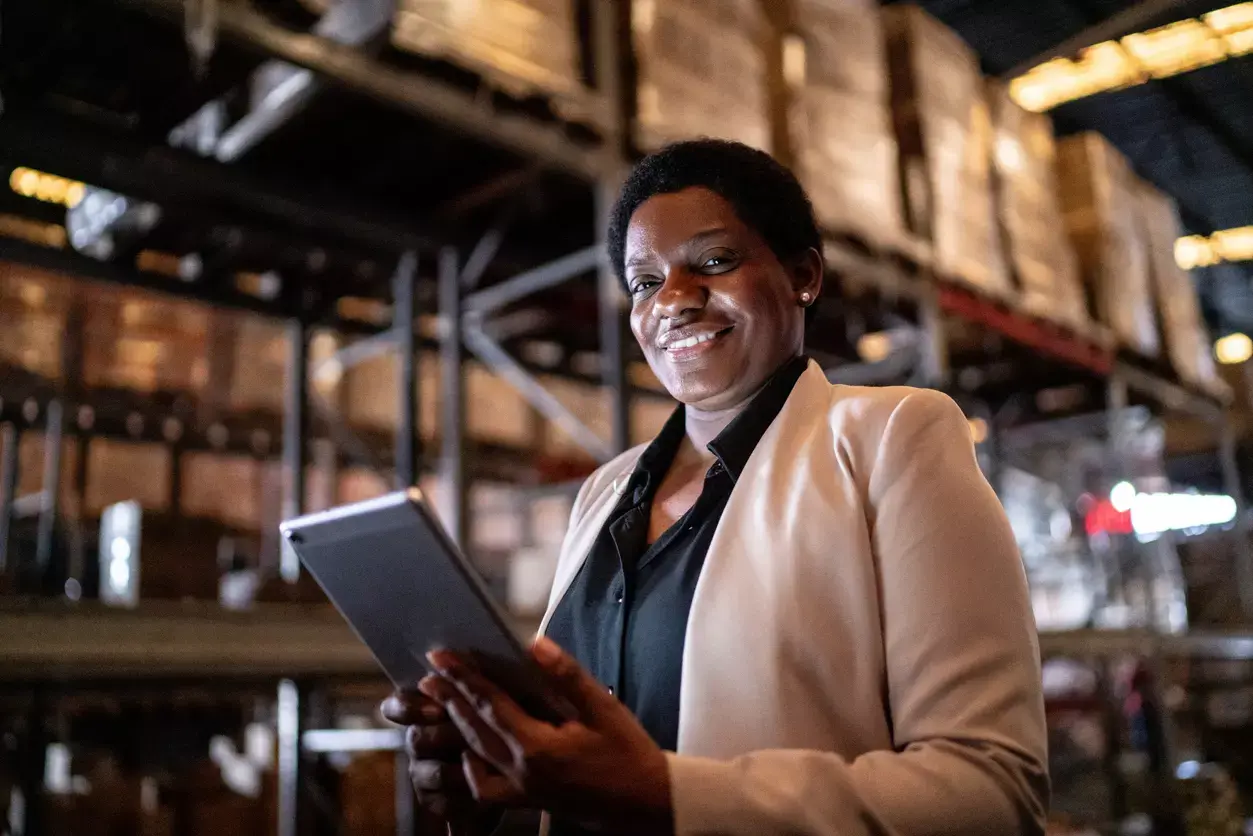 Smiling businesswoman using tablet in warehouse to apply for a loan, shelves of inventory visible behind her.