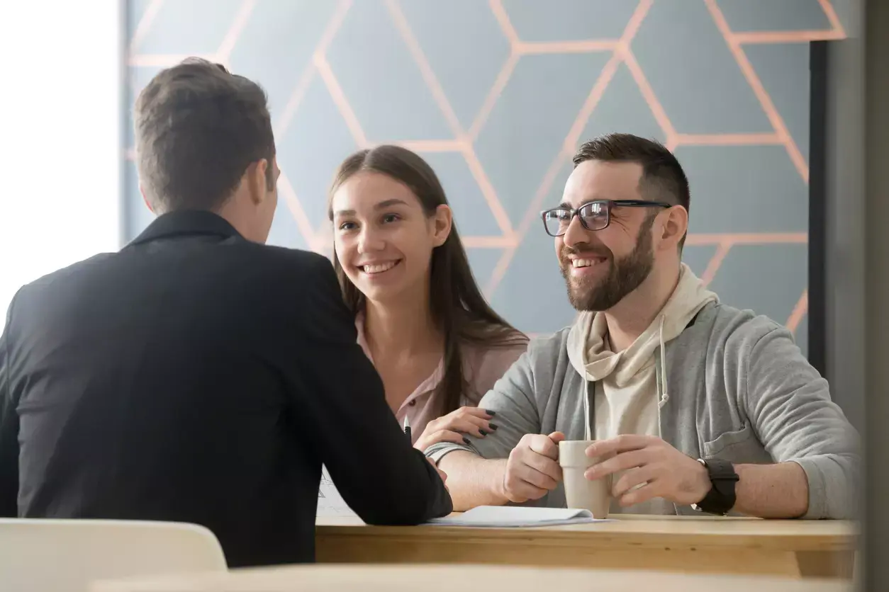 Smiling professionals discuss business loans at modern office table. Geometric wall pattern in background.