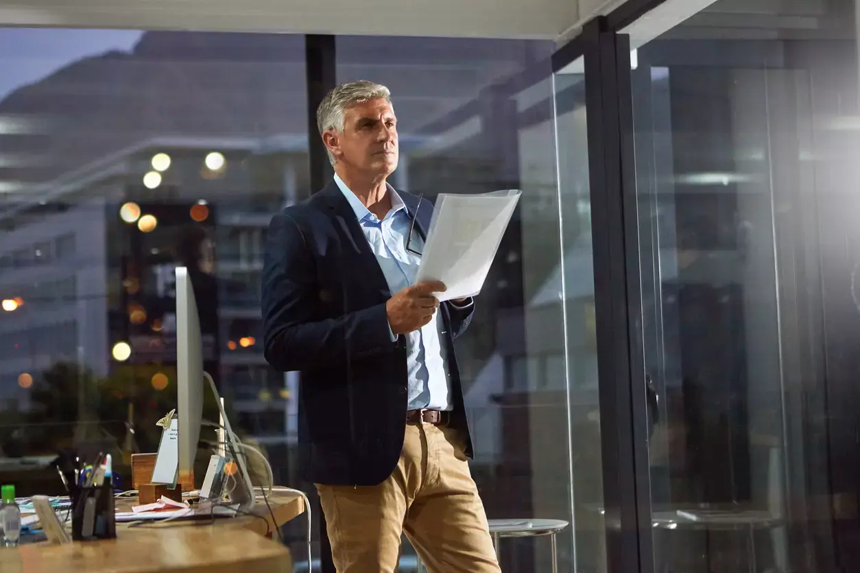 Mature businessman examines loan application in modern office, city lights visible through windows at dusk.
