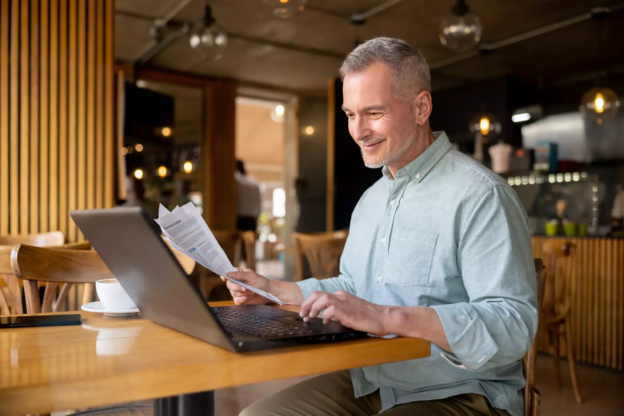 Smiling older man in cafe analyzes documents while working on laptop, reviewing debt documentation