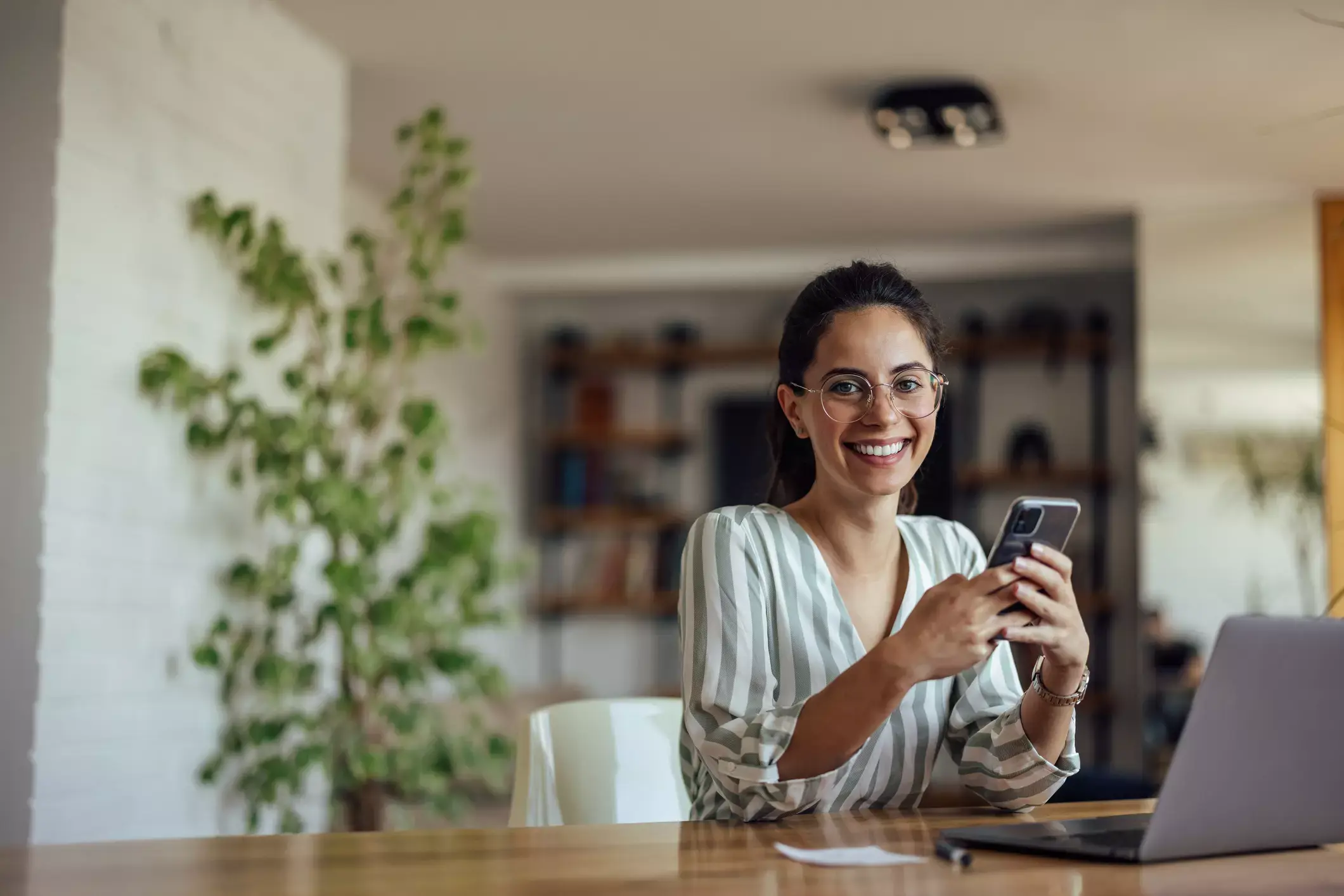 Smiling professional woman in striped blouse using smartphone at home office desk with laptop and plant nearby