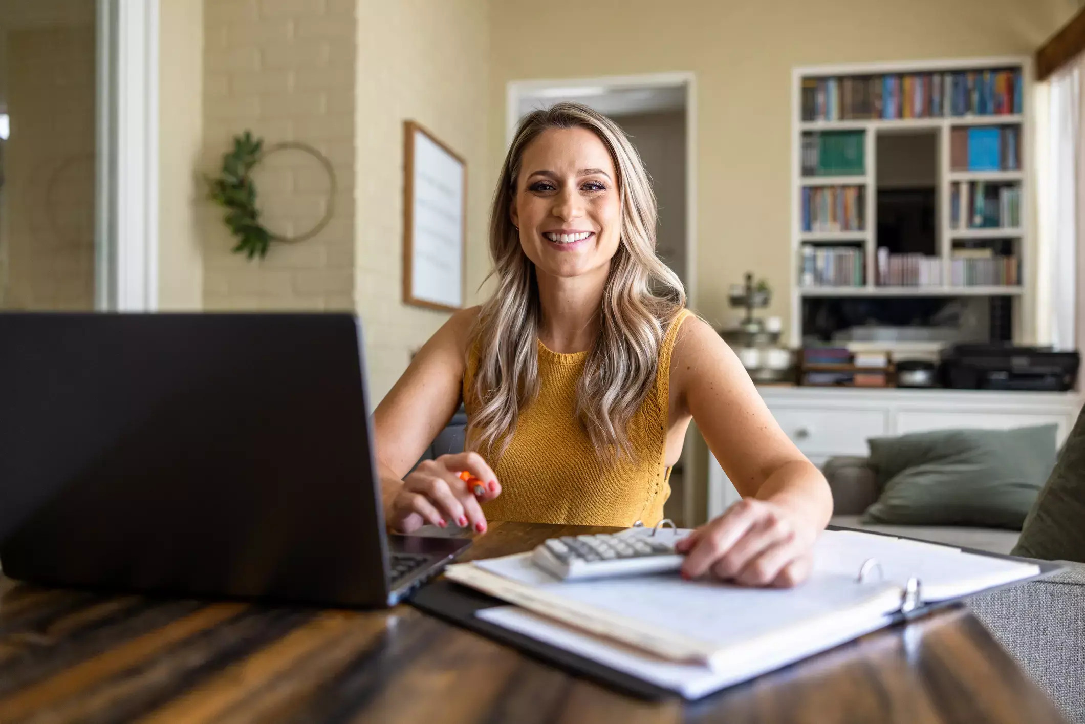 Smiling woman in yellow top using laptop and calculator, reviewing business loan paperwork in home office