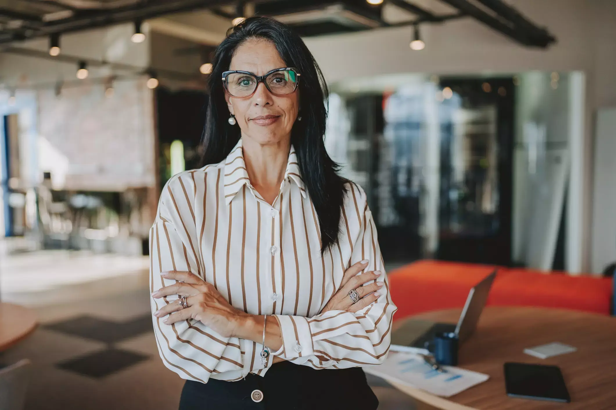 Mature professional woman in striped shirt stands confidently in modern office, arms crossed, ready to advise entrepreneurs