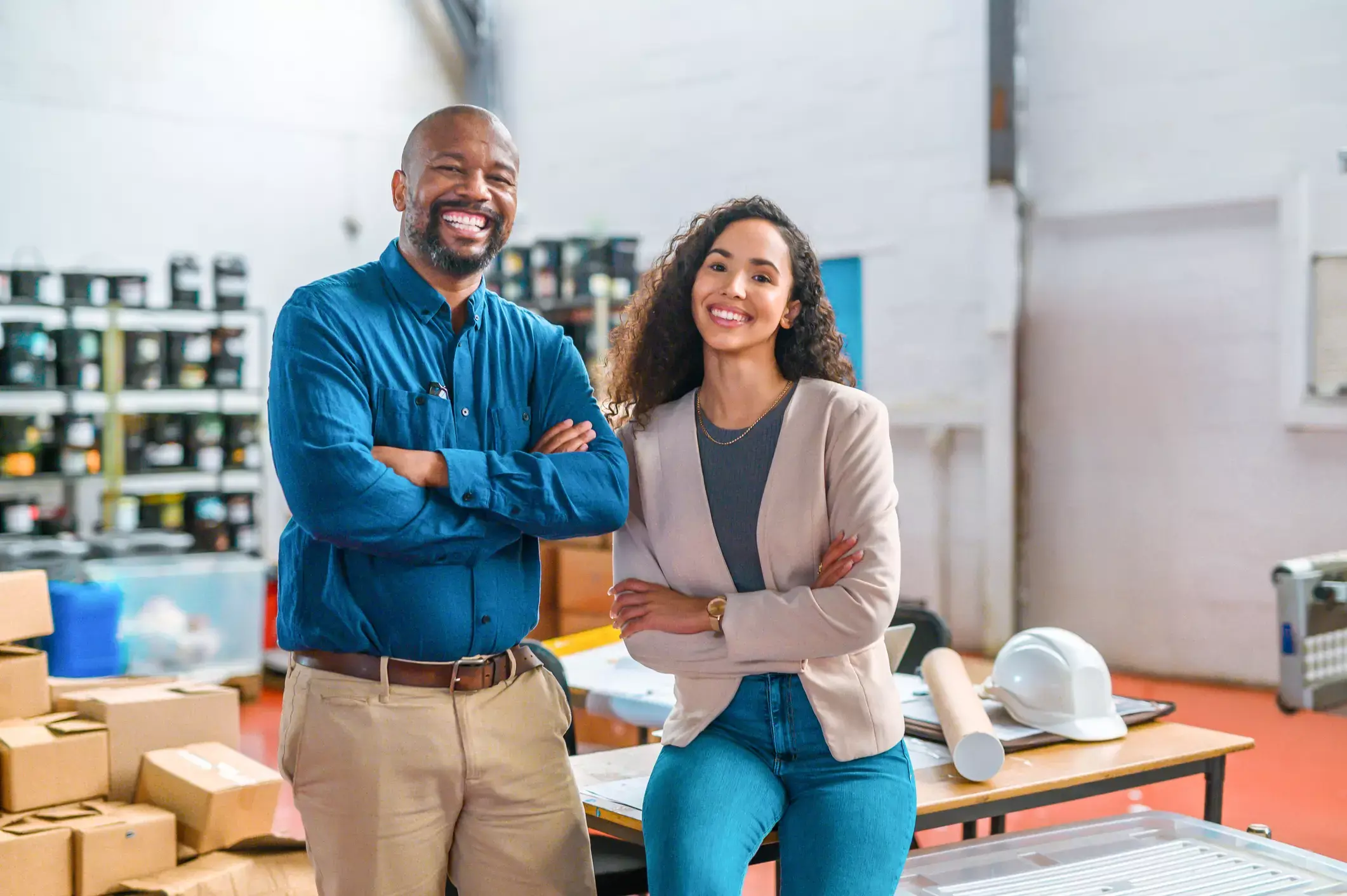 Smiling African-American man and Hispanic woman, business partners, standing confidently in warehouse with supplies and equipment.
