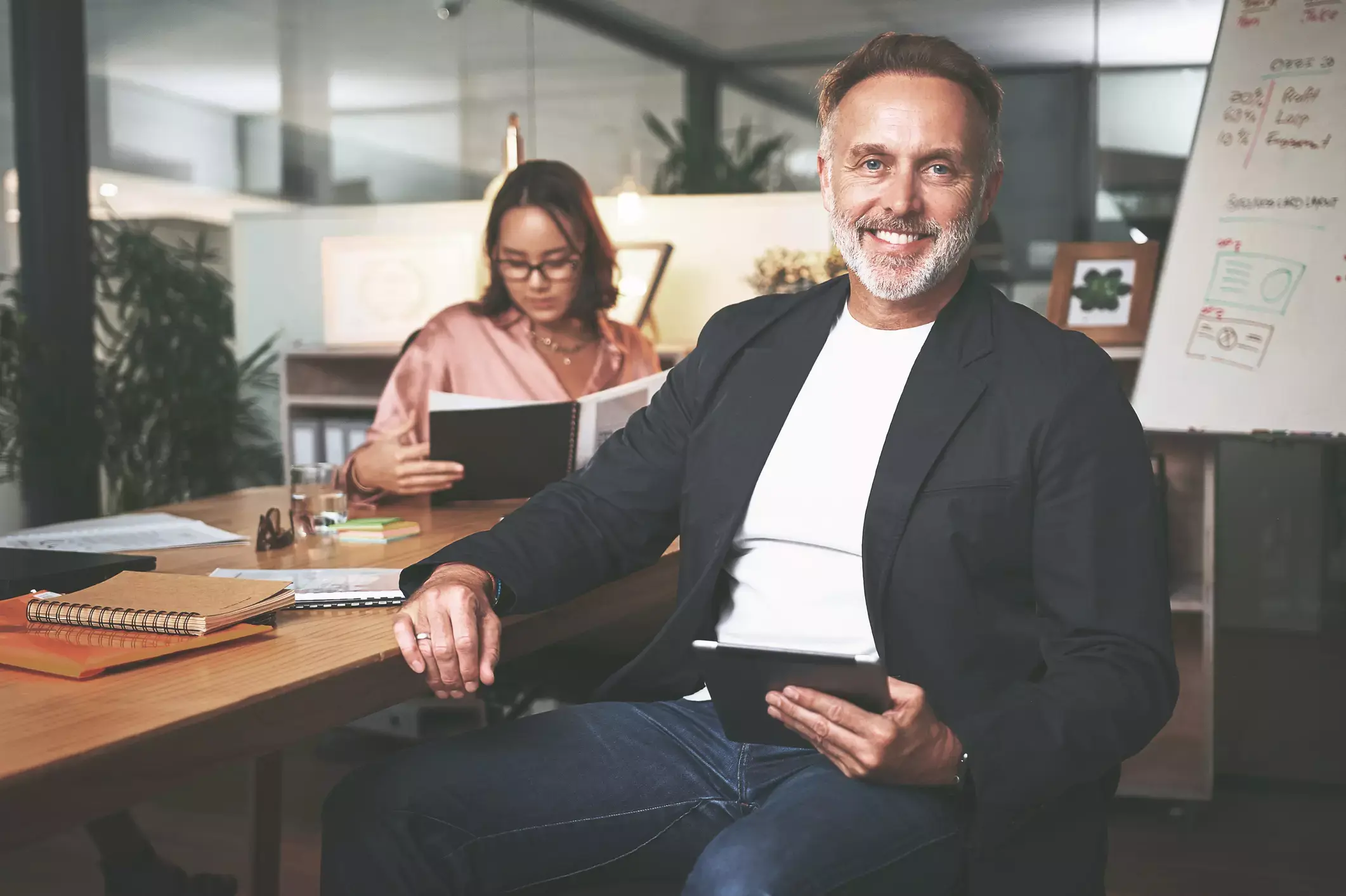 Senior businessman smiles confidently, holding tablet, as young woman reviews loan application in modern office workspace